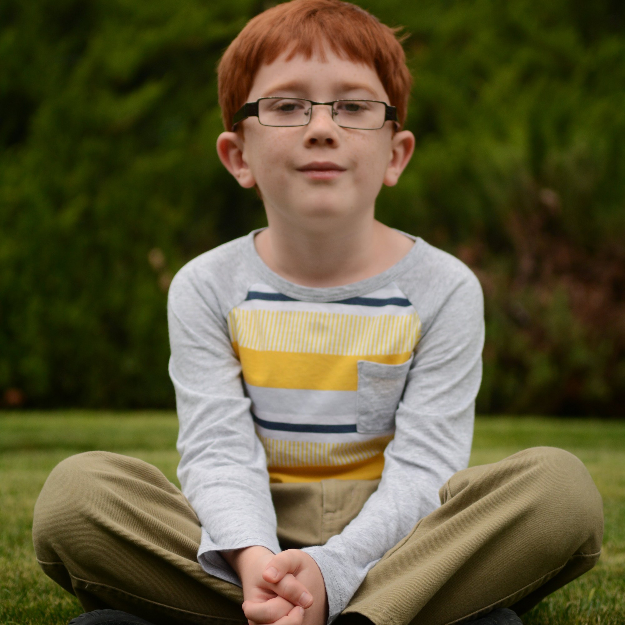A young boy sits on a grassy lawn, wearing a long-sleeved gray shirt with yellow and white stripes, tan pants, and black s...