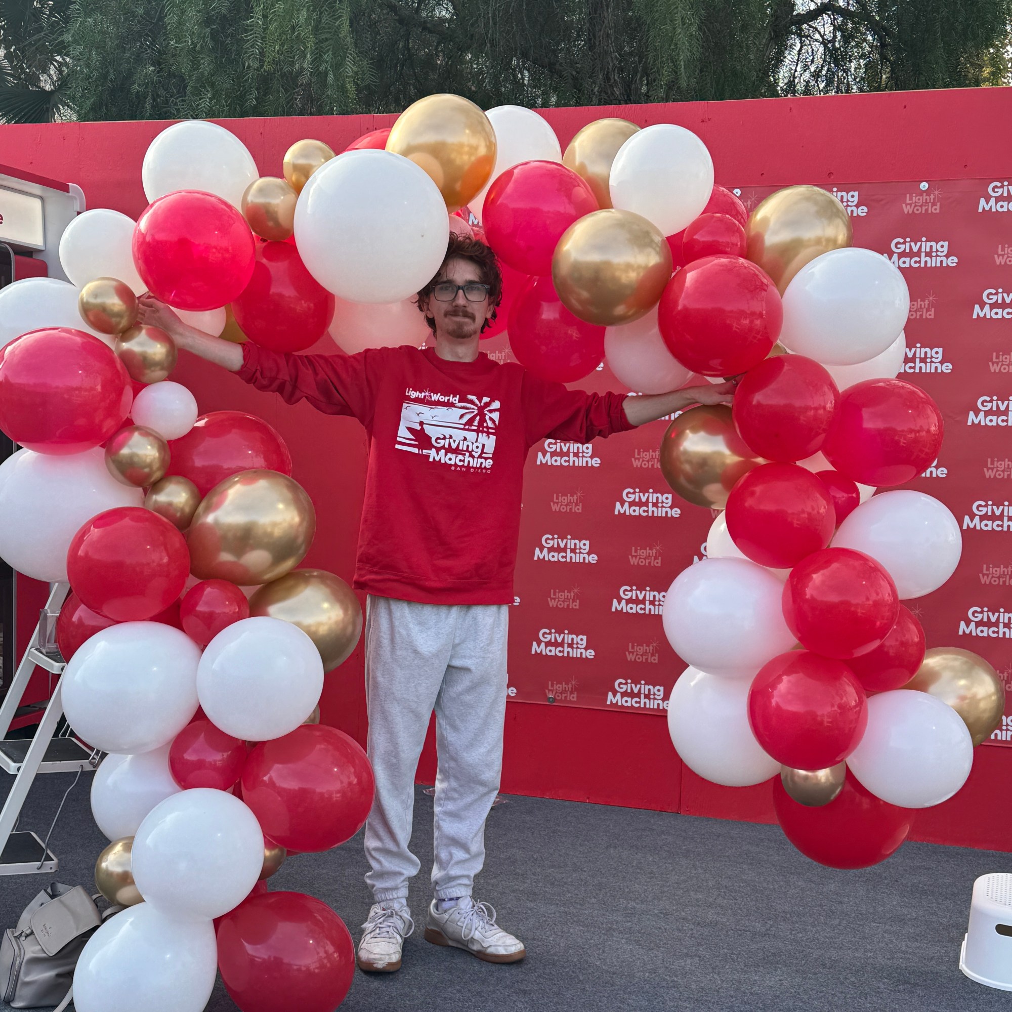 A man stands proudly in front of a vibrant red backdrop, donning a red sweatshirt emblazoned with "Light the World" and "G...