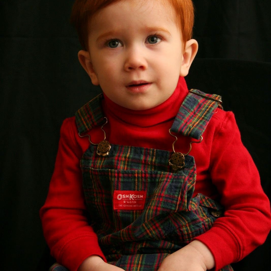 A Red-haired Child in Osh Kosh B'Gosh Overalls Sitting in Front of a Black Background.
