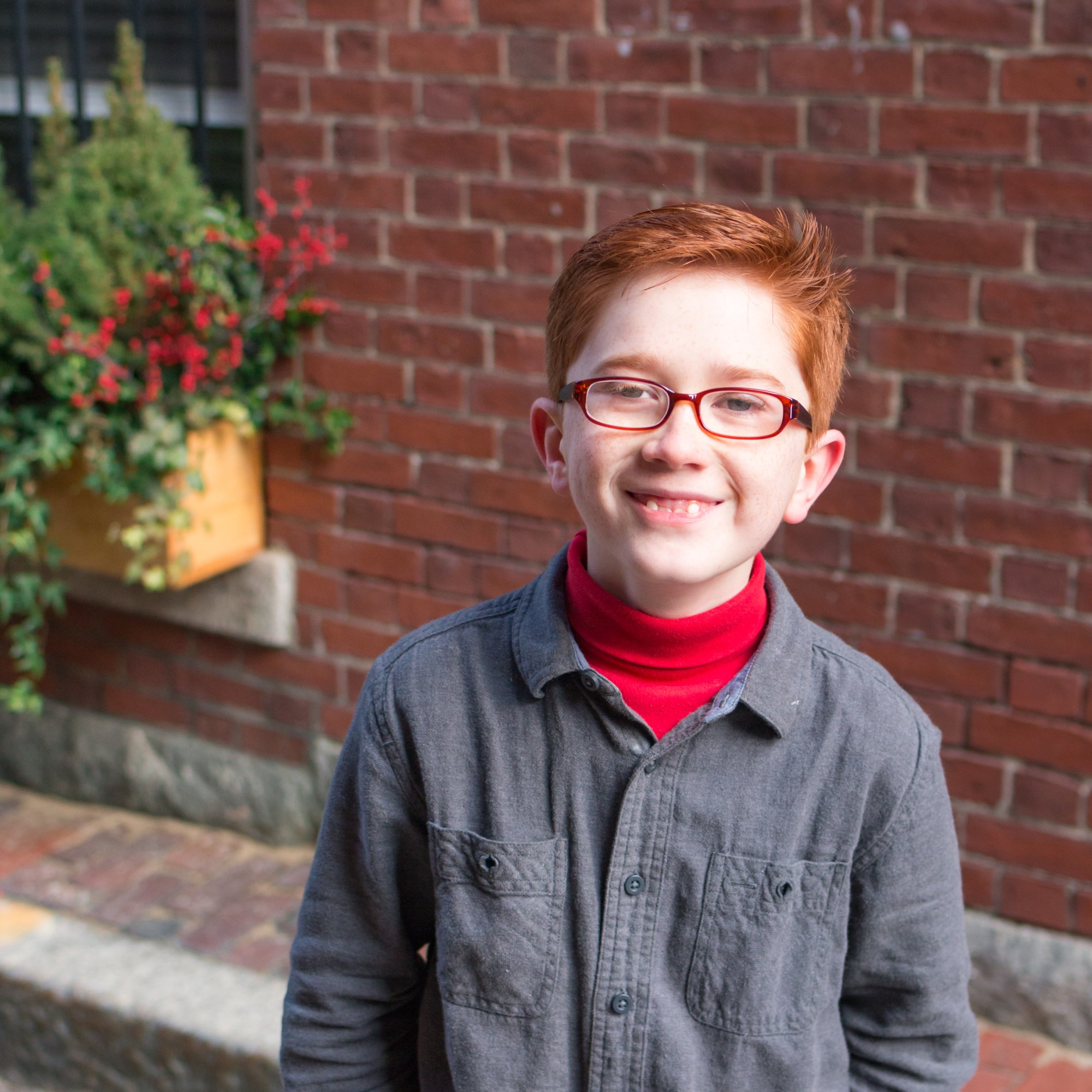Here is the alt text: "boy with red freckles, red glasses, and shirt, in front of brick wall."