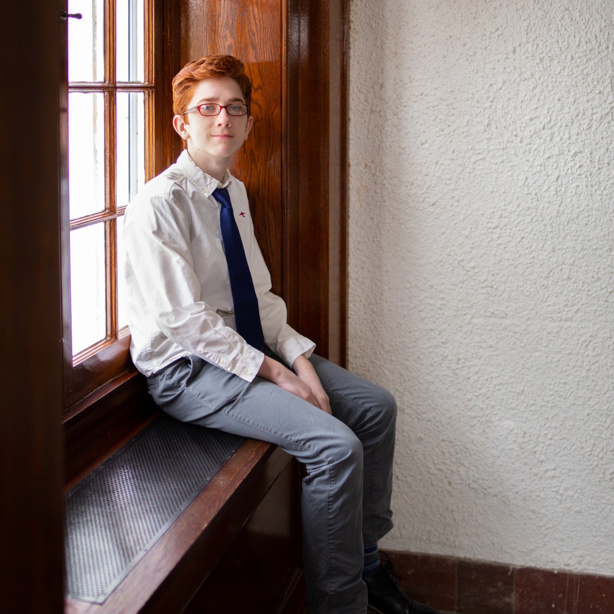 A young person with red hair and glasses sitting on a window-seat ledge. The background is a white wall.